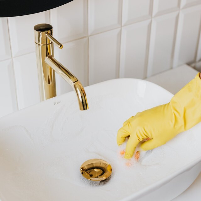 Gloved hand cleaning a white bathroom sink with a gold faucet